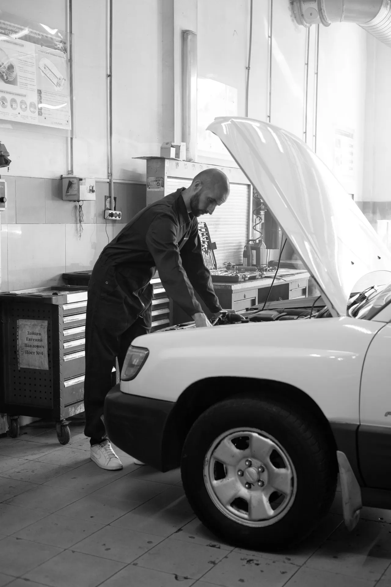 Illustrative black-and-white photograph of a mechanic inspecting a vehicle under its open hood